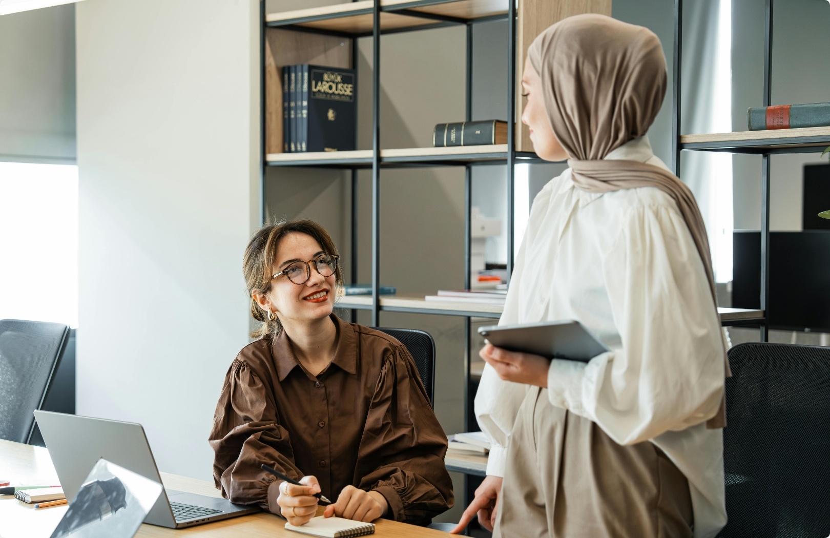 Eine Muslima und eine europäisch gelesene Frau unterhalten asich am Schriebtsich in einem Büro